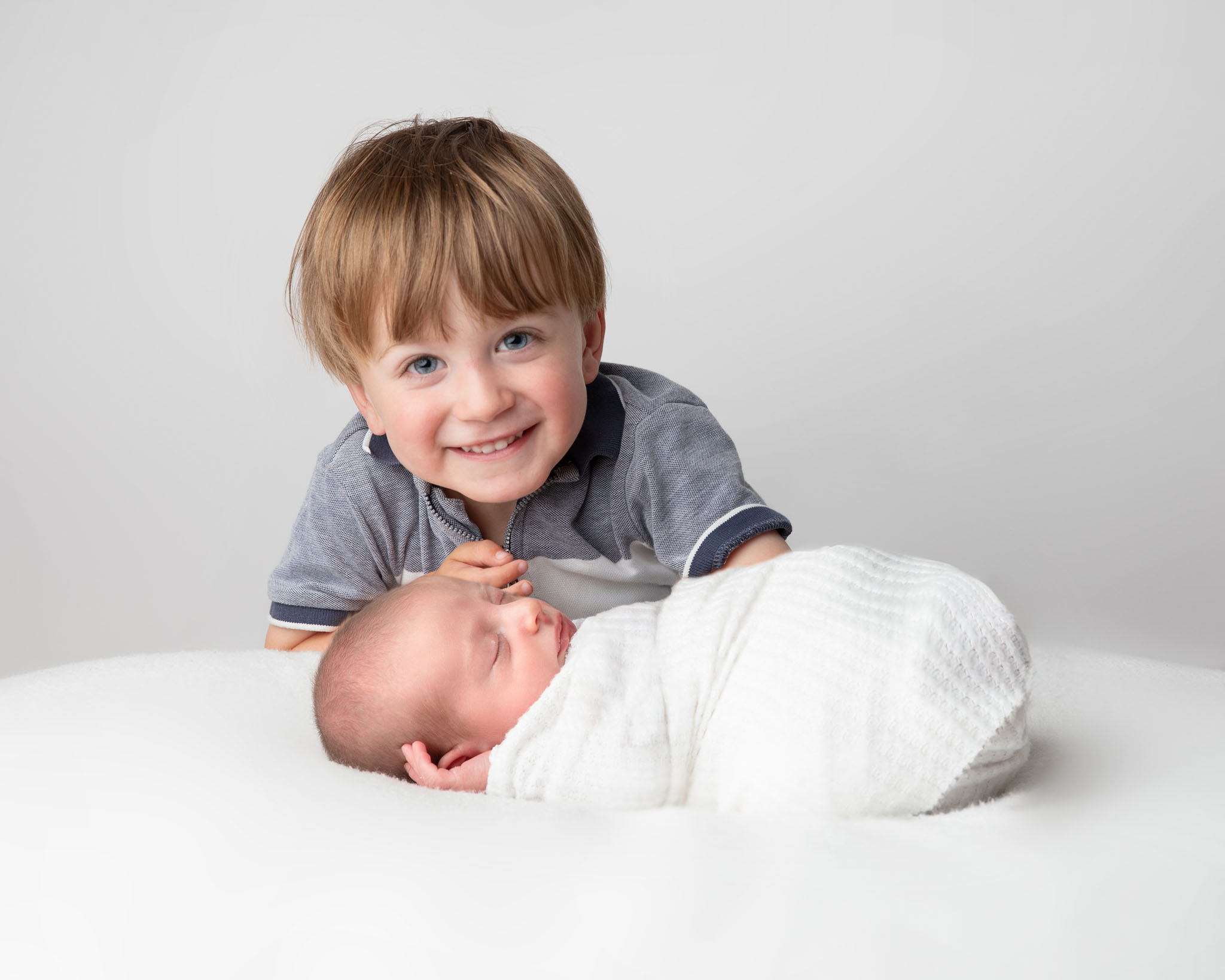 A proud older brother smiling at the camera while he looks down on his new baby by Newborn photographer in Somerset, Wiltshire and Bath