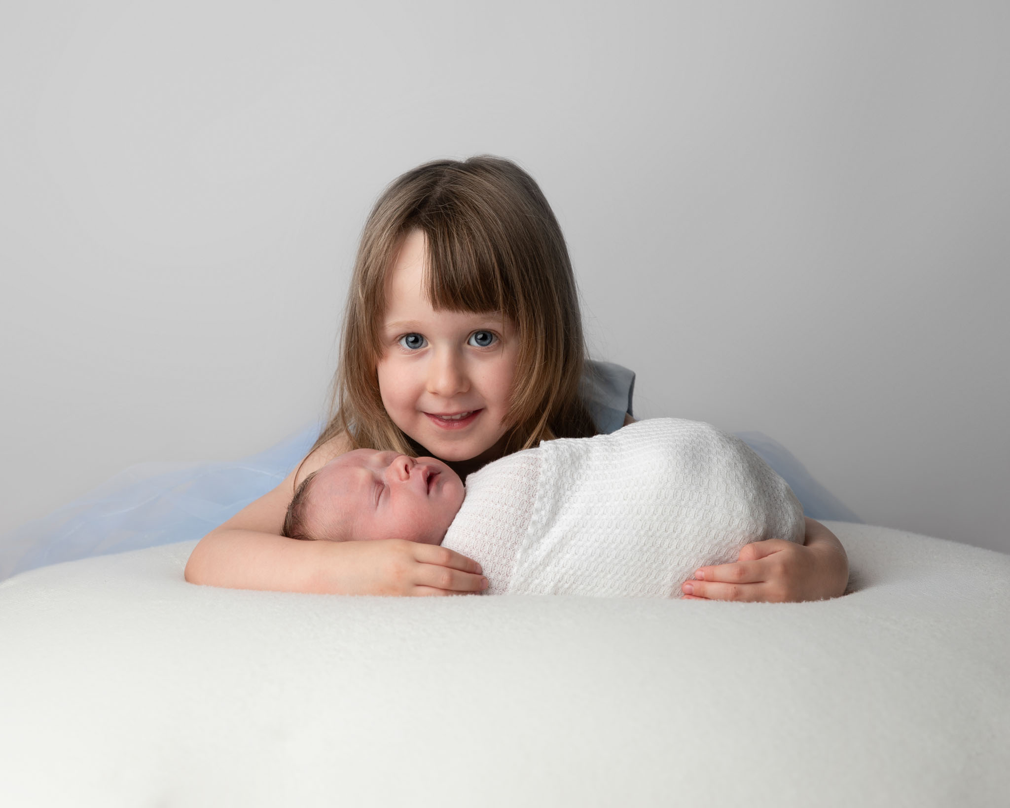 proud older sister holds her newborn baby brother on a white beanbag in a photography studio by Newborn photographer in Somerset, Wiltshire and Bath<br />
